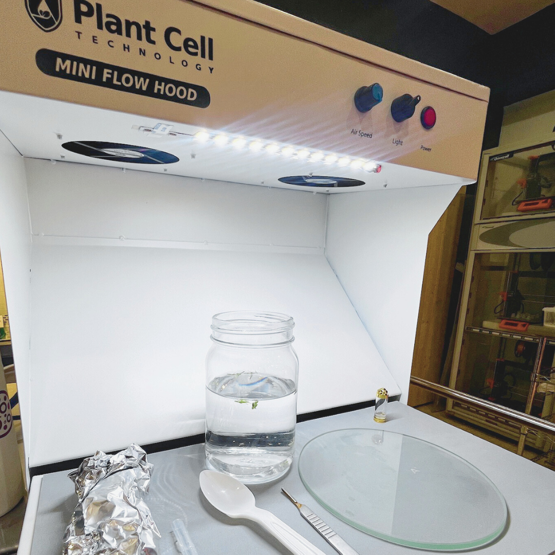 Discover 'The Mini' portable flow hood by Plant Cell Technology. The image showcases floating plants in jar, tissue culture sample jewelry, glass plates, and various tools.